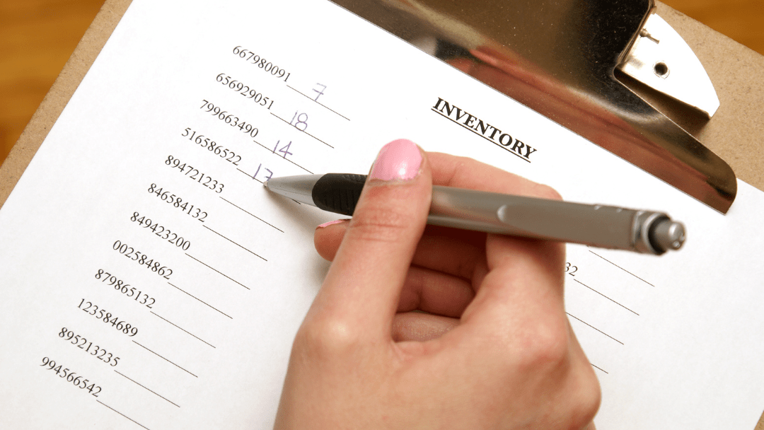 A hand holding a pen and writing numbers on a printed inventory sheet clipped to a clipboard, illustrating manual inventory management.